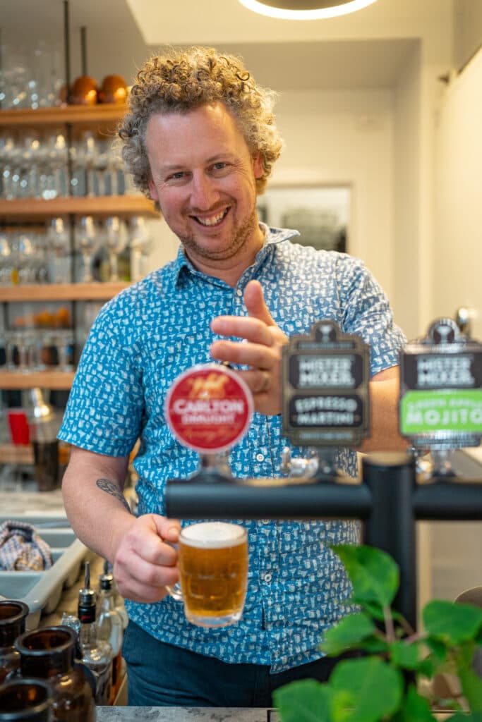 A smiling man holding a glass of beer behind a bar