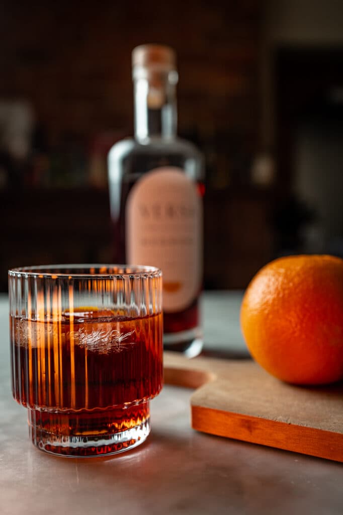 An amber cocktail with a large ice cube beside an orange