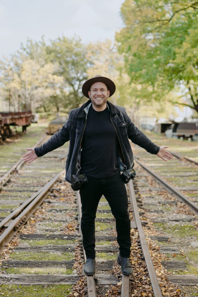 A smiling man in a hat standing on railway tracks